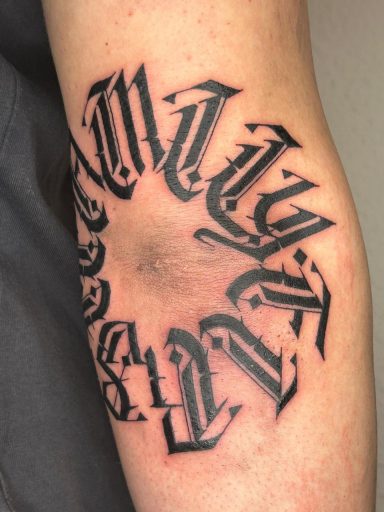 Schwarzes Tattoo mit gedrehten Buchstaben in kreativer Schriftart auf dem Unterarm.