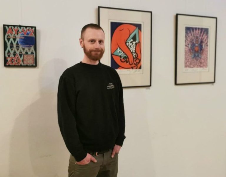 Mann steht neben bunten Kunstwerken an einer Wand in einem Ausstellungsraum.