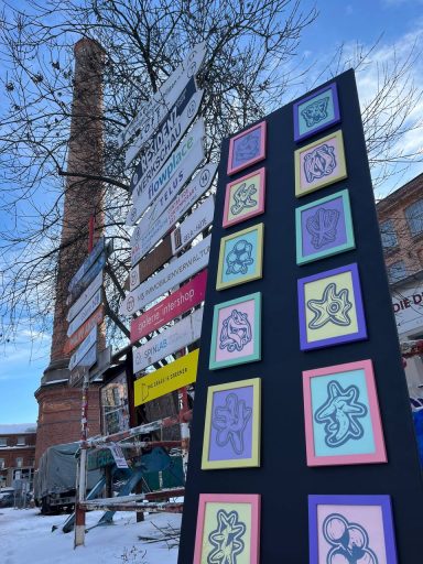 Bunte Kunstwerke an einem schwarzen Schild vor einem alten Schornstein im Schnee.