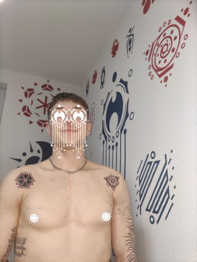 Mann mit Tattoos auf dem Oberkörper, vor einer bunten Wand mit grafischen Mustern.